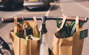 Two brown paper bags with handles and with plants placed inside hanged on a bicycle's handlebar while on the center of the street.